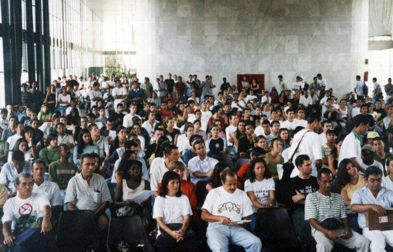 Auditório de paredes brancas e cadeiras pretas, quase todas lotadas. À esquerda da foto, grandes janelas de vidro por onde entra a luz do sol.