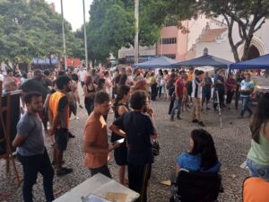 Dezenas de pessoas de pé, reunidas em círculo, aguardam o início do evento. Algumas estão de colete laranja, identificando a Asav Sindicato. Ao centro, um adulto jovem manipula uma câmera sobre um tripé. 