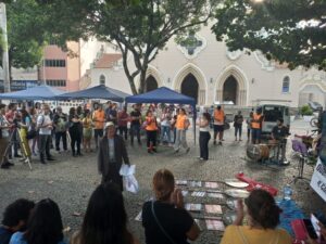 Dezenas de pessoas de pé, reunidas em círculo ao redor de cartazes e placas organizadas no chão, observam a fala de uma mulher jovem de colete laranja. Ao fundo está uma igreja bege com três grandes portas abertas. 