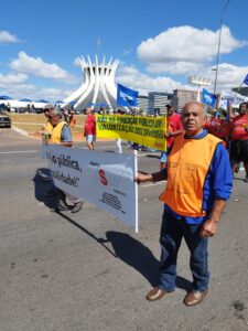 Tem homens idosos caminham por uma avenida próxima à catedral, da direita para a esquerda, segurando uma longa faixa com o logotipo da Asav. Eles usam coletes laranjas. 