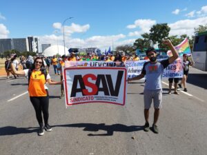 Um homem e uma mulher jovens seguram uma faixa com o logotipo da Asav e caminham numa avenida larga na nossa direção. Eles estão bom os punhos fechados e os braços erguidos. Atrás deles, Muitas outras pessoas caminham na nossa direção, segurando várias faixas. 