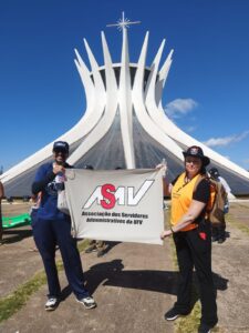 Dia claro sem nuvens no céu. Pátio amplo de cimento com uma grande catedral em formato de cone ao fundo, formada por colunas brancas que se se encontram no topo. Em frente a ela, um homem e uma mulher posam para a foto segurando uma bandeira com o logotipo da Asav. 