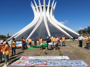 Dia claro sem nuvens no céu. Pátio amplo de cimento com uma grande catedral em formato de cone ao fundo, formada por colunas brancas que se se encontram no topo. Em frente a ela, várias faixas estendidas no chão com nomes de sindicatos e palavras de ordem. Várias pessoas transitam por entre as faixas, algumas delas tocando tambores e outros instrumentos de percussão. 