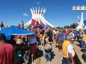 Pátio amplo de cimento com catedral em forma de cone ao fundo. Muitas pessoas, de variados gêneros, idades e raças caminham pelo local, alguns segurando bandeiras com nomes de sindicatos. 