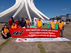 Dia claro sem nuvens no céu. Pátio amplo de cimento com uma grande catedral em formato de cone ao fundo, formada por colunas brancas que se se encontram no topo. Em frente à catedral, um grupo de pessoas posa para a foto segurando uma longa faixa vermelha onde se lê: Asav pela reposição salarial, por verbas para a Universidade Pública e contra a retirada de direitos. 