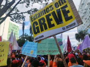 Grande número de pessoas de pé estão próximas a um prédio verde de 4 andares. Muitas delas seguram placas, faixas e cartazes com os dizeres "greve", "estamos em greve" e outros acima de suas cabeças. 