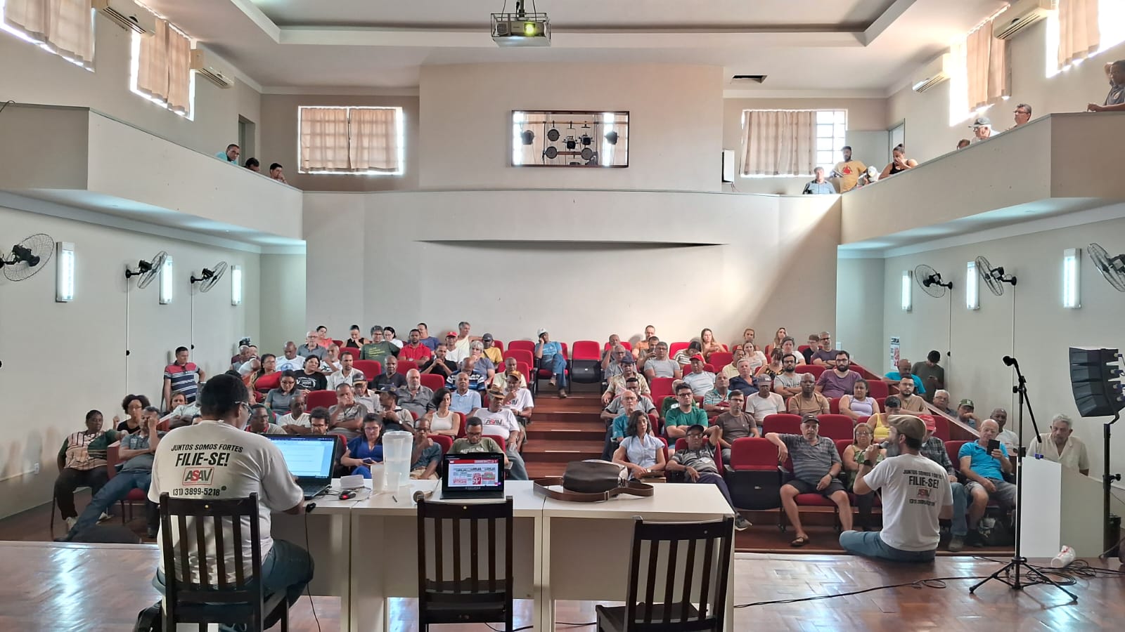 Foto tirada do fundo do palco em direção à plateia. Em primeiro plano, há uma mesa branca com um homem negro de camiseta branca sentado em uma cadeira, de costas para nós. Ao fundo, as pessoas da plateia estão sentadas em cadeiras vermelhas, de frente para nós. à direita, um homem de boina e camisa branca está sentado na beirada do palco, de costas para nós, e parece falar o microfone para a plateia. No alto das paredes laterais direita e esquerda do auditório há varandas voltadas para dentro com uma fileira de pessoas em cada uma, assistindo à assembleia. 