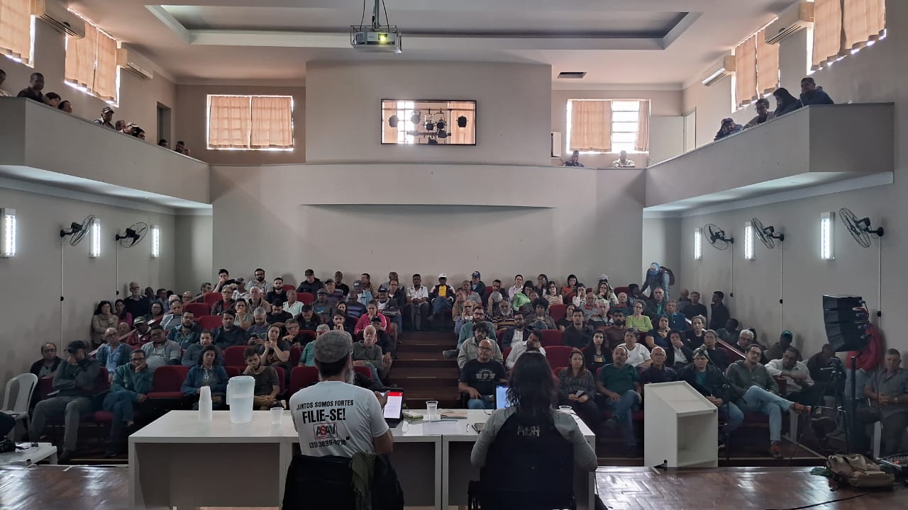 Auditório branco com todas as cadeiras ocupadas. Nas laterais, há balcões a cerca de 3 metros de altura, com uma fileira de pessoas em cada um. Sobre o palco de piso de madeira, há uma mesa branca com um homem e uma mulher sentados a ela. A foto foi tirada por detrás destes dois, do fundo do palco.