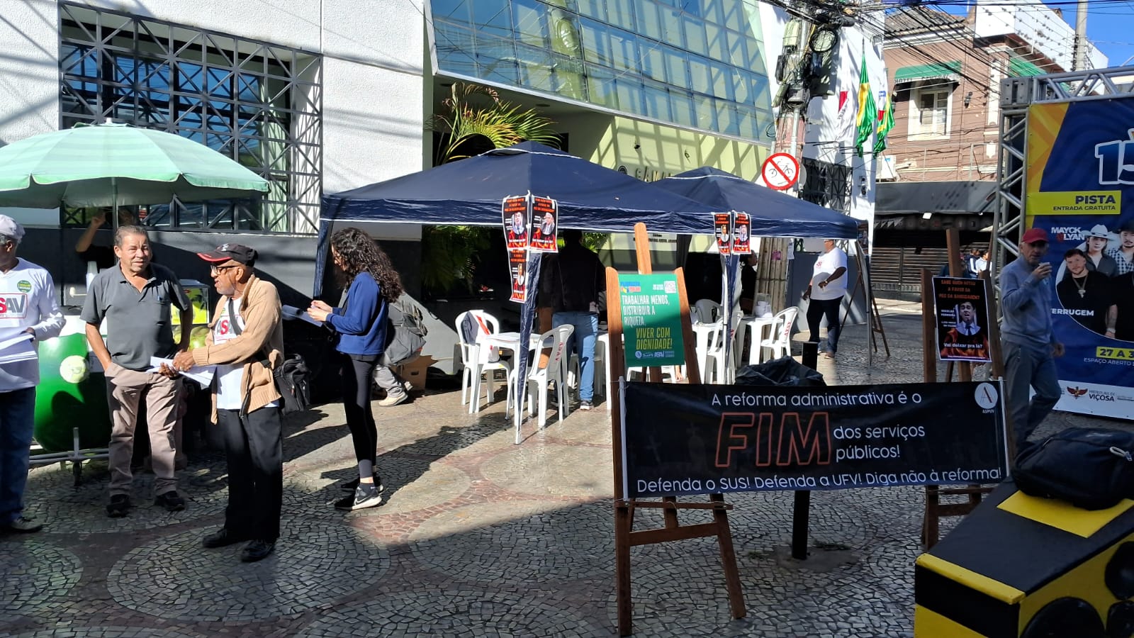 Foto afastada mostra as duas tendas azuis ao centro; à esquerda um carrinho de venda de côco gelado com um guarda-sol verde claro. Próximo de nós há uma faixa presa entre dois cavaletes de madeira onde se lê "A reforma administrativa é o FIM dos serviços públicos. Defenda o SUS! Defenda a UFV! Diga NÃO à reforma administrativa!". Há um pequeno movimento de pessoas ao redor. 