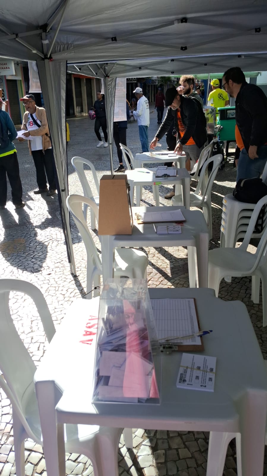 Foto tirada debaixo de uma das tendas, mostrando a fileira de mesas e cadeiras plásticas brancas, com urnas de votação e papéis sobre elas. Há um pequeno movimento de pessoas ao redor. 
