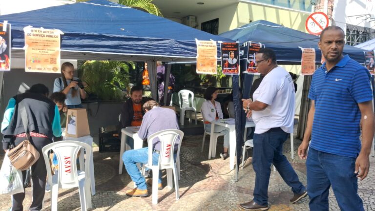 Duas tendas azuis grandes, montadas lado a lado. Sob elas há várias mesas e cadeiras plásticas brancas, com pessoas sentadas atrás delas e algumas sentadas à frente. Sobre as mesas há urnas de madeira e de plástico transparente, por onde se vê vários papeis dentro. Um homem negro de camisa branca caminha da direita para a esquerda em frente a uma das tendas. Há uma grande movimentação de pessoas ao redor.