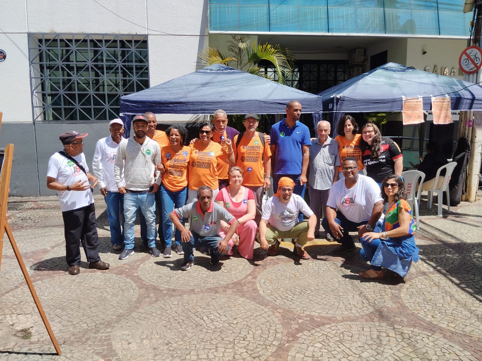 11 homens e 6 mulheres, de diferentes etnias e idades, lado a lado e olhando na nossa direção. Parte destas pessoas está de pé e a outra parte está agachada em frente a eles. Atrás do grupo, há duas grandes tendas azuis com cartazes colados nelas. 