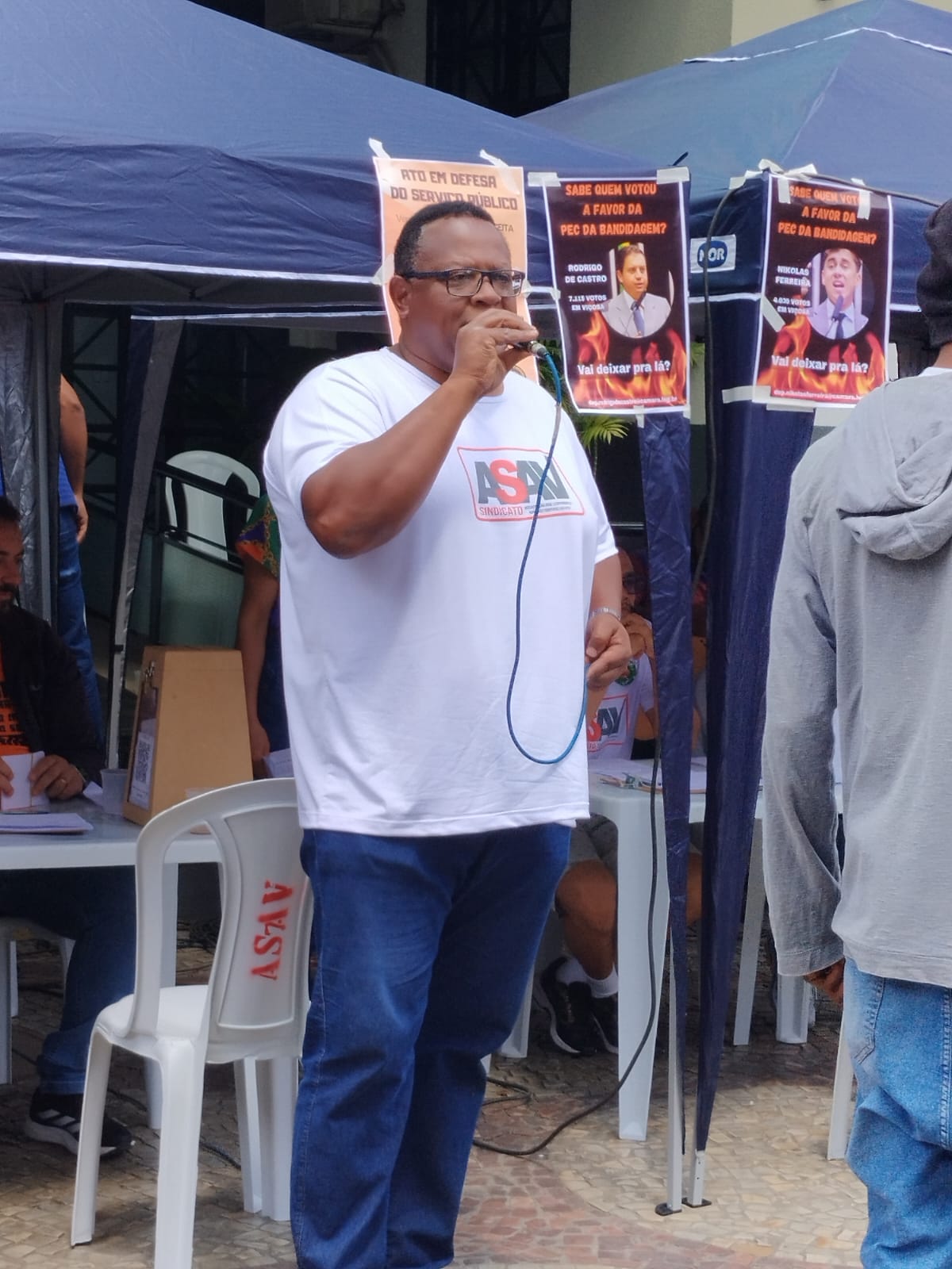 Homem negro robusto, de óculos corretivo, e camisa branca com a logo da Asav. Ele está de pé virado para a direita e segura um microfone em frente à boca com a mão esquerda. 