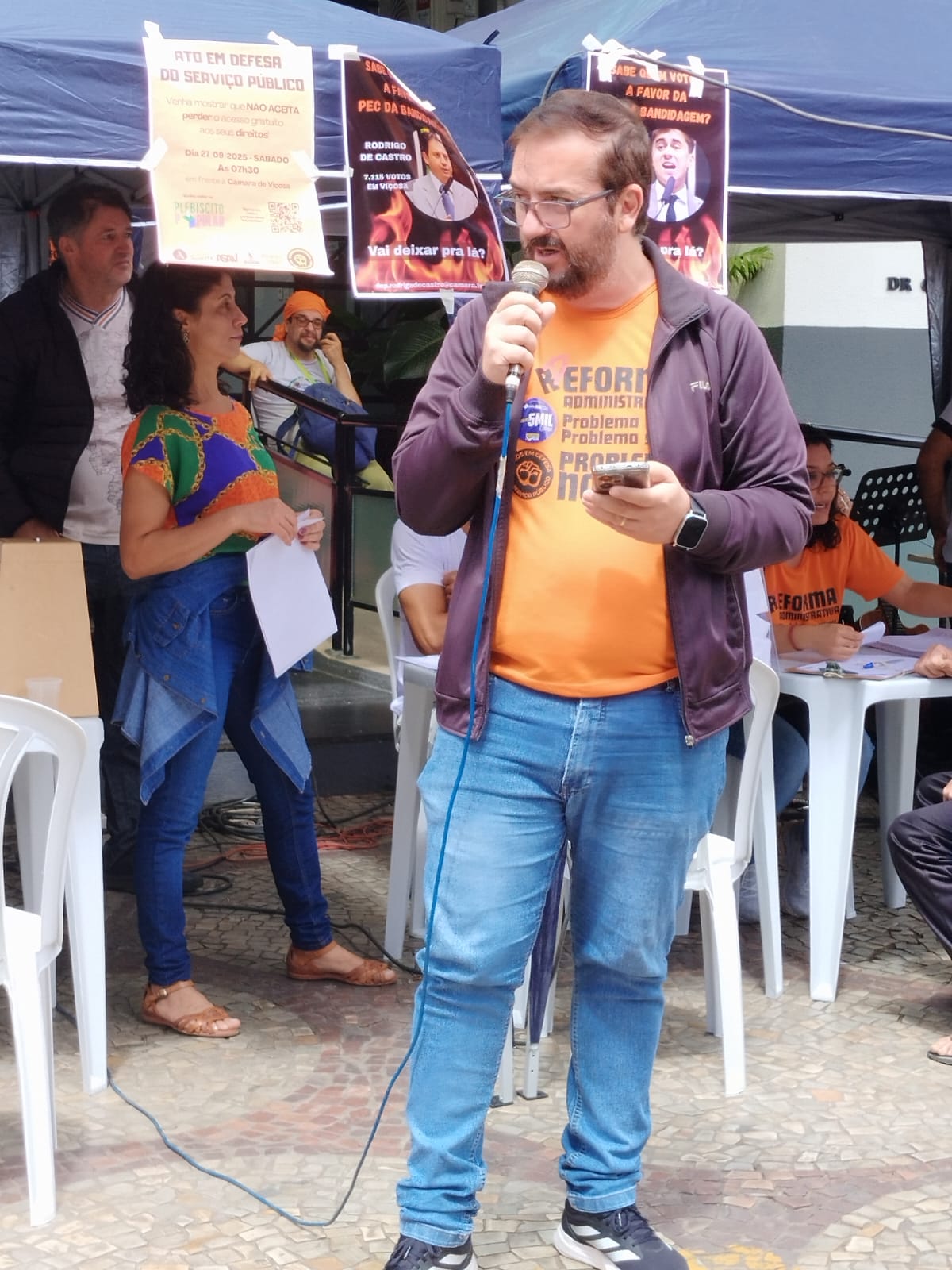 Homem branco alto, de camiseta laranja e casaco roxo, óculos corretivo, cabelo barba e bigode castanho claros. Ele está de pé olhando para a esquerda, segurando um microfone em frente à boca com a mão esquerda e um celular com a direita. 