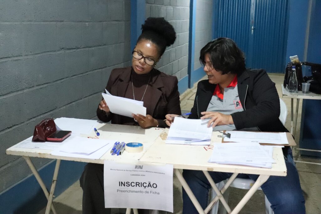 Edimara e Aline sentadas atrás de duas mesas metálicas brancas. Edimara é uma mulher negra, magra, cabelos crespos presos no alto da cabeça e óculos; ela examina alguns papéis. Aline é uma mulher parda, cabelos pretos ondulados na altura do queixo e óculos. Ela se inclina na direção de Edimara e parece falar. 