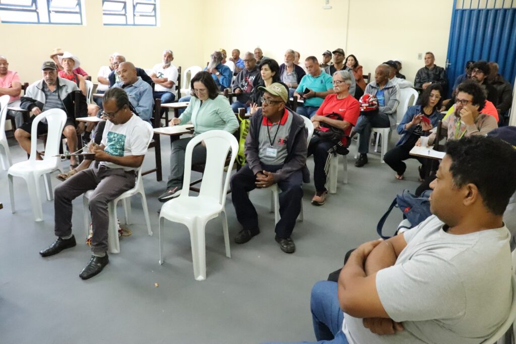 Sala de reuniões amarelo claro com várias cadeiras de madeira e de plástico branco, quase todas ocupadas por pessoas idosas, ms também há pessoas mais jovens. O piso é de cimento cinza e a porta é de metal ondulado azul escuro. 