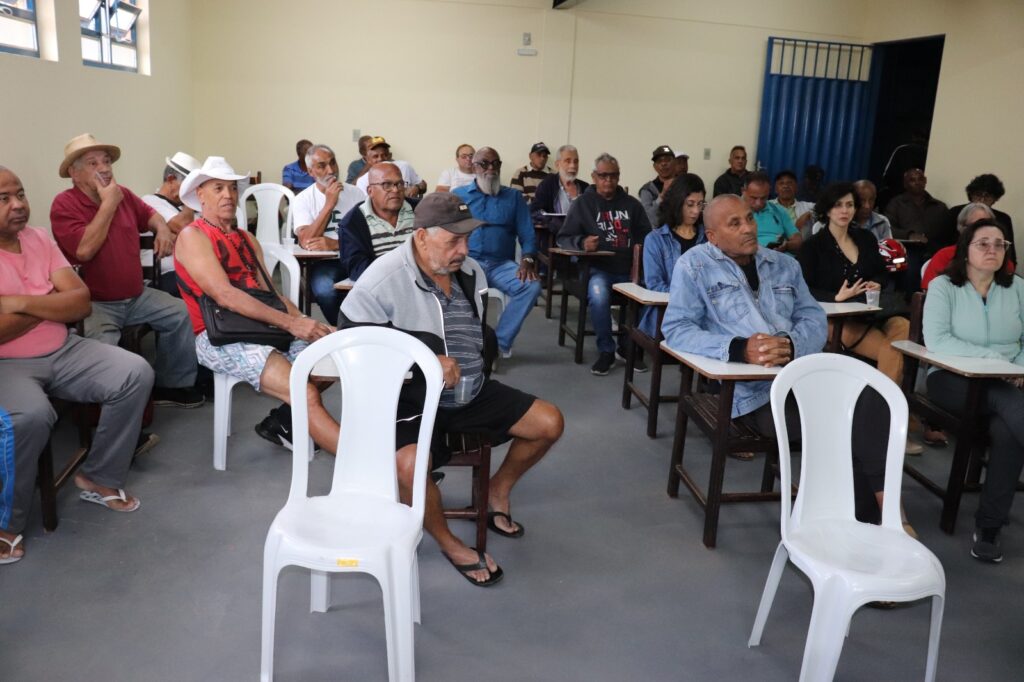 Sala amarelo claro com várias cadeiras de madeira e de plástico branco, quase todas ocupadas, em sua maioria, por homens idosos. Homens e mulheres mais jovens também estão presentes. Três homens estão de chapéu branco. 
