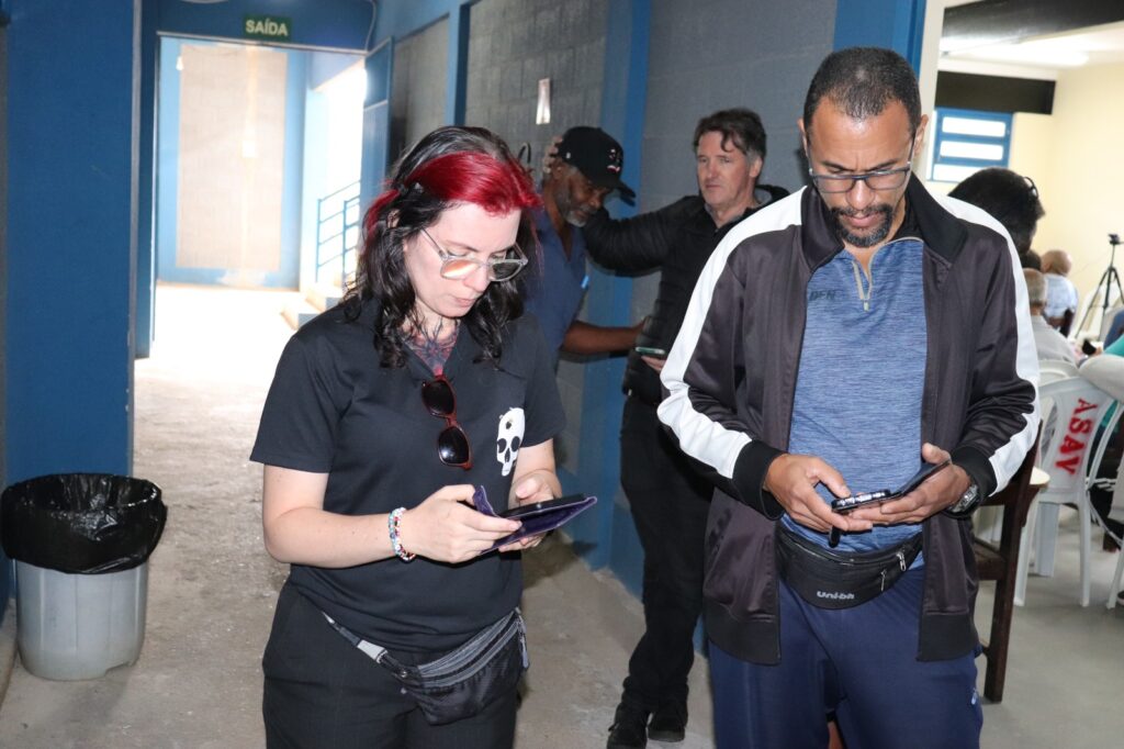 Verônica e Rodrigo de pé no corredor em frente à sala de reuniões, ambos mexendo no celular. Verônica é uma mulher jovem branca e magra, cabelos ondulados longos com uma grande mecha na franja pintada de vermelho; ela usa óculos e roupas pretas. Rorigo é um homem pardo de meia-idade, ligeiramente calvo, barba e cavanhaque pretos levemente grisalhos; ele também usa óculos. 