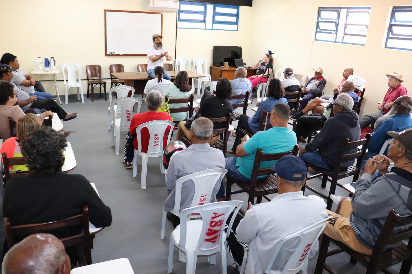 Giovani, homem branco magro de meia-idade, boina bege, barba longa e bigode, fala sentado em uma mesa. Em frente a ele, uma plateia formada principalmente por homens idosos olha em sua direção.