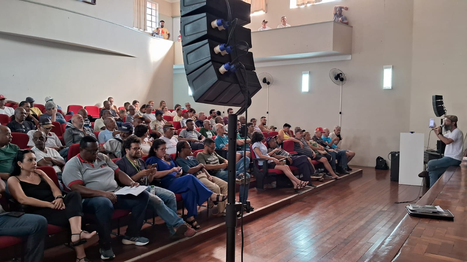 Auditório de paredes brancas e piso de madeira. À esquerda, fileiras de cadeiras vermelhas, quase todas ocupadas por pessoas de variados gêneros, idades e raças. À direita, sentado no palco, há um homem de camisa branca, barba longa, bigode e usando boina falando ao microfone, olhando na direção da plateia. 