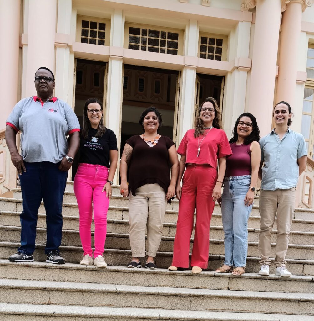 Grupo formado por dois homens e 4 mulheres, todos de pé lado a lado, no degrau de uma escadaria do Prédio Principal da UFV.