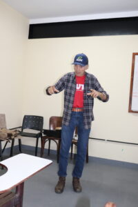 Saulo: homem idoso branco, alto, magro, cabelos grisalhos sobressaindo ao boné, camiseta vermelha com camisa cinza de mangas longas por cima. Ele está de pé com os braços erguidos na altura do peito e fala para os presentes. 