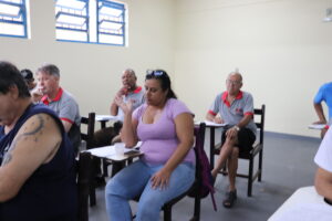 Renata: mulher de meia-idade parda, robusta, cabelos pretos lisos e longos, óculos no alto da cabeça e camiseta roxa. 