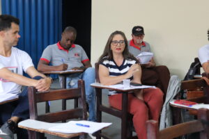 Aloirta: mulher de meia-idade branca cabelos castanhos longos, óculos corretivo e batom vermelho. À esquerda dela, Rodrigo Cardoso: homem jovem branco, alto, magro, cabelos castanhos rentes à cabeça e camiseta branca. Atrás de Aloirta estão Cleves (homem de meia-idade negro, magro, camiseta cinza e olhando para baixo enquanto anota em uma folha) e um homem idoso branco, de boné preto, bigode e cavanhaque brancos. 
