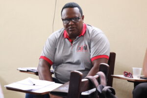 Evaristo: homem de meia-idade negro, robusto, cabelos pretos rentes à cabeça, óculos corretivo, camisa cinza de gola vermelha. 