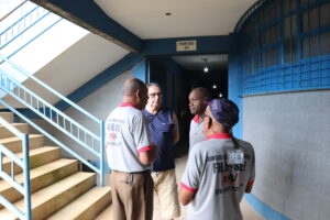 4 homens idosos de diferentes estaturas e raças, de pé próximos a uma escada com corrimão azul claro. 