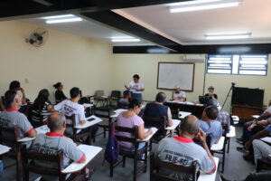 Sala de paredes amarelo claro com dezenas de cadeiras de braço de madeira, a maioria delas ocupada por pessoas de diferentes idades , gêneros e raças. Há um quadro branco na parede à frente, e também uma mesa com um homem de boina e uma mulher de cabelos curtos. Uma mulher está falando, de pé, perto da mesa.