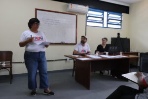 Aline de pé ao lado da mesa fazendo uma fala. Ela é uma mulher parda de meia-idade, robusta, cabelos pretos curtos e lisos, óculos corretivos e camiseta branca. À mesa estão Giovani (homem de meia-idade branco, magro, usando boina e óculos corretivos) e Beatriz (mulher de meia-idade branca, cabelos grisalhos curtos e cacheados, óculos corretivos e camiseta preta. 
