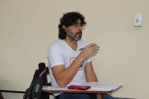 Mairréuli: homem de meia-idade branco, alto, magro, cabelos pretos cacheados e volumosos, bigode e barba ligeiramente grisalhos, óculos corretivos e camiseta branca. Ele está com a boca ligeiramente aberta e a mão esquerda erguida em frente ao corpo. 