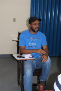 Manoel: homem jovem negro, magro, alto, óculos corretivo, bigode e barba pretos, boné preto e camiseta azul. 