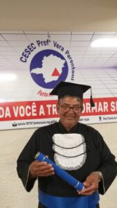 Homem idoso, negro, cabelos grisalhos e óculos. Ele sorri e usa roupa e chapéu de formatura e segura um canudo de formatura azul escuro. 