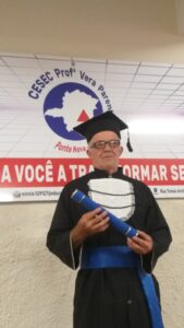 Homem idoso, branco, cabelos grisalhos e óculos. Ele usa roupa e chapéu de formatura e segura um canudo de formatura azul escuro. 