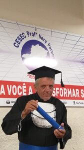 Homem idoso, pardo, cabelos grisalhos, olhos caídos e ombros ligeiramente encolhidos para cima. Ele usa roupa e chapéu de formatura e segura um canudo de formatura azul escuro. 