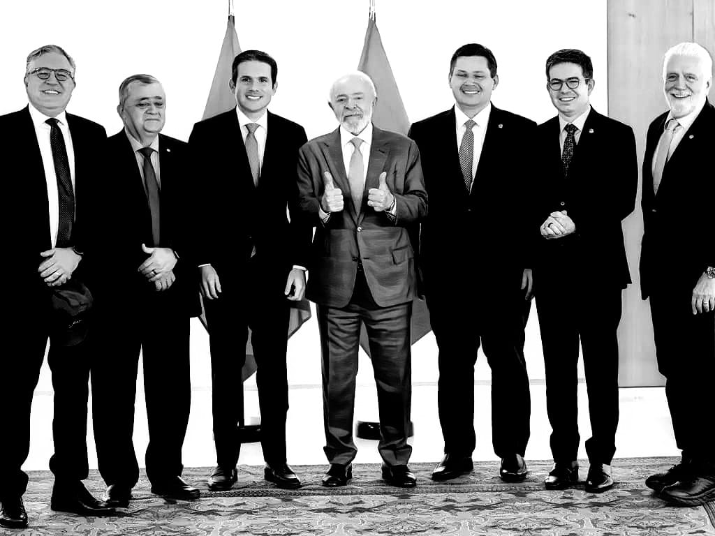 Foto em preto e branco. 7 políticos de pé, lado a lado, todos de terno e gravata, olhando na nossa direção e sorrindo. O do meio é o presidente Lula, que faz "joinha" com as duas mãos.