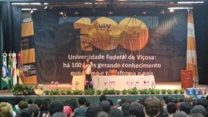 Palco de auditório com um grande cartaz ao fundo com a logo do centenário da UFV. No centro dele, uma mesa decorada com toalha branca. Em frente à mesa, a deputada Duda Salabert: mulher, alta, cabelos longos castanhos e ondulados, óculos corretivos, calça preta e camisa branca. 