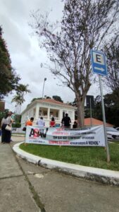 Faixa com a logo da Asav instalada em gramado em frente à casa onde funciona a reitoria da UFV. Algumas pessoas estão de pé pelo local, e outra s estão de pé na varanda da reitoria. O tempo está nublado. 
