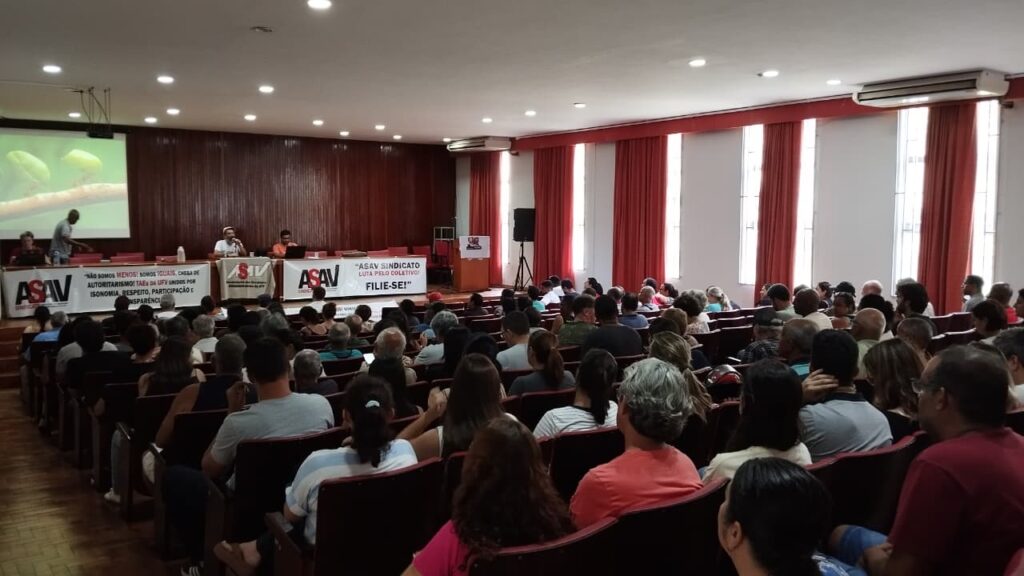 Assembleia quase totalmente lotada, num auditório com janelas altas parcialmente cobertas com cortinas vermelhas. Em frente à mesa há várias faixas com a logo da ASAV Sindicato. 