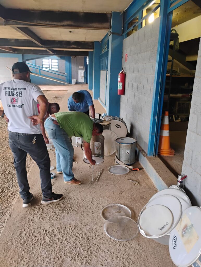 4 homens em frente a um galpão coberto e piso sem acabamento. Perto deles há várias peças de tambores desmontados. Três desses homens estão mexendo com algumas das peças no chão. 