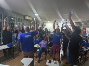 Dezenas de pessoas de diferentes idades, gêneros e etnias, a maioria de pé. Elas estão erguendo um crachá azul no ar. Pelas laterais de vidro do salão, percebe-se que já é noite. 