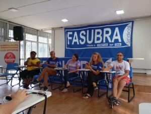 Grupo de 5 pessoas de diferentes idades, etnias e gêneros sentados lado a lado. Uma mulher negra fala ao microfone. Na parede ao fundo, há uma grande bandeira azul com a logo da Fasubra. 
