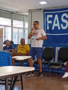 Homem jovem, magro, negro, usando óculos, bermuda azul e camiseta da Asav está de pé falando ao microfone, perto de outras duas pessoas sentadas. 