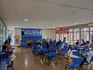 Salão com piso de madeira e paredes de vidro com grades brancas. Há dezenas de cadeiras azuis, quase todas ocupadas. Em frente a estas cadeiras, um grupo de 5 pessoas está sentado de frente para a audiência. 