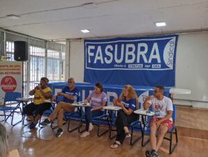 5 pessoas de diferentes idades, gêneros e etnias, todas sentadas. Uma mulher negra fala ao microfone, e as demais a observam. 