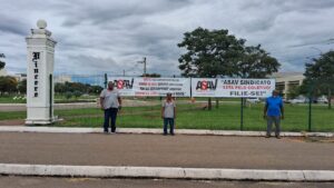 Faixas com a logo da ASAV Sindicato presas em uma grade, próximo a uma pilastra com a palavra "vincere". Três homens negros estão de pé em frente às faixas olhando na nossa direção; dois deles usam camisa cinza com a logo da Asav, e o outro usa camisa azul. 
