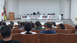 Foto de auditório com algumas cadeiras ocupadas. Ao fundo, uma longa mesa coberta por faixas com a logo da Asav e chamados para filiação. Um homem negro robusto, de camisa cinza, está sentado no centro e falando ao microfone. 