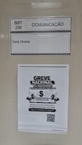 Cartazes fixados numa porta bege. O de cima tem uma tabela com o nome da BBT 230 Comunicação; o de baixo traz o texto Greve Nacional dos TAEs, ASAV. 